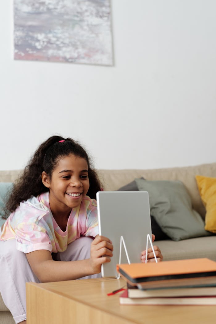 Smiling teenager using a tablet for online learning at home, sitting comfortably with books and a cozy couch.